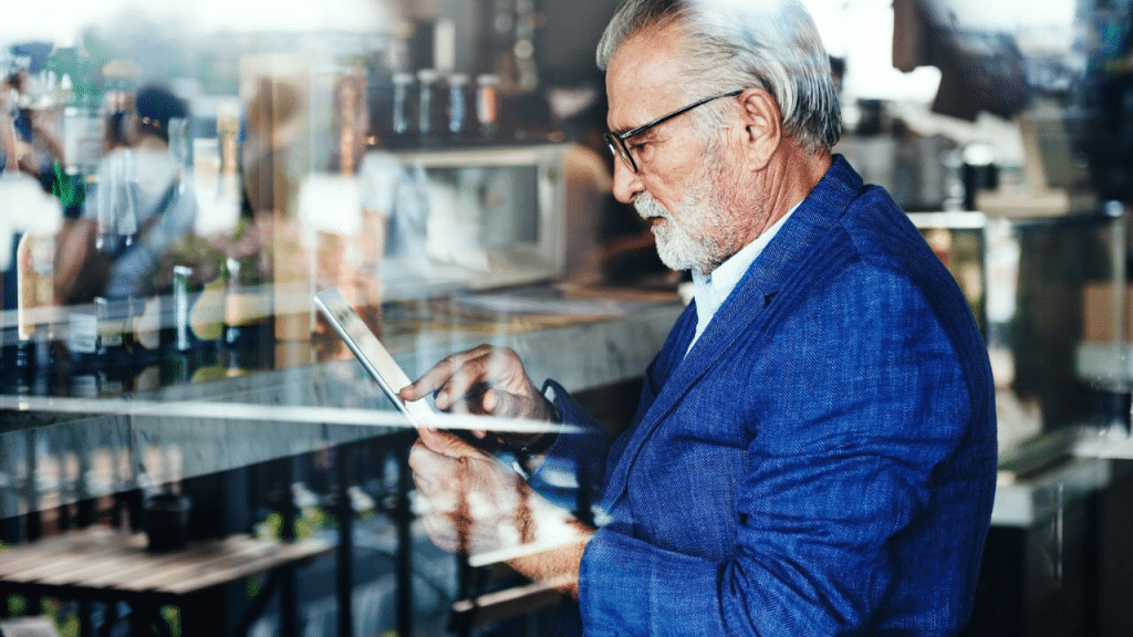 A senior businessman in a blue blazer and glasses is seated at a table and using a tablet.