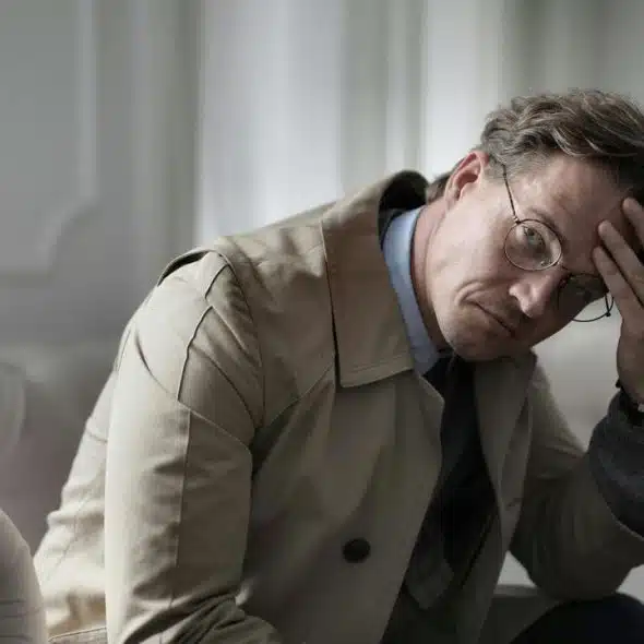 A man in glasses and a trench coat sitting on a couch with his hand on his forehead.