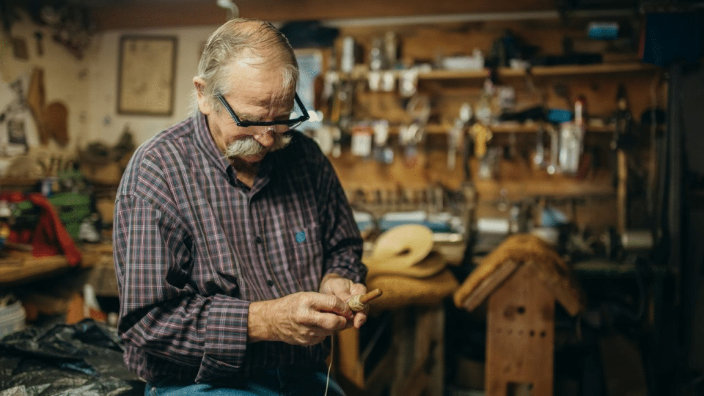 A thoughtful, senior Caucasian man with a mustache and glasses is seated in a workshop and working on a small object.