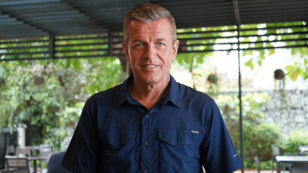 A smiling middle-aged man in a blue collared shirt is standing outdoors and looking at the camera.