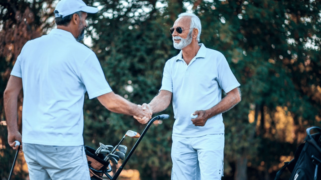 A handsome, older man with a beard and sunglasses is shaking hands with another man in an all-white golf outfit.