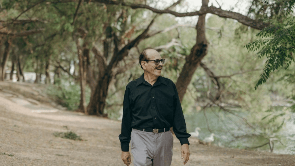 A smiling older man with a mustache and sunglasses is walking on a path in a park.