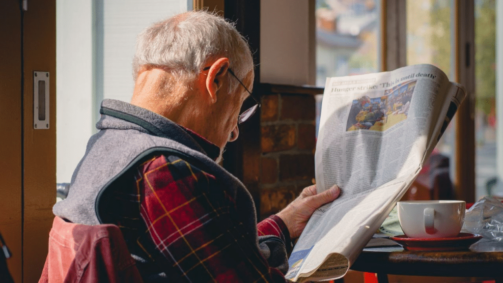 A senior man in glasses is seated at a table, holding a newspaper with both hands and reading.
