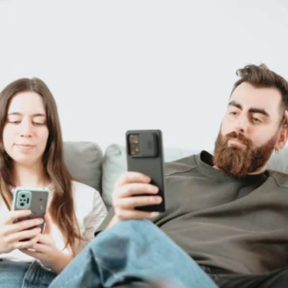 A man and woman sitting together in a living room, both looking distant while holding their phones