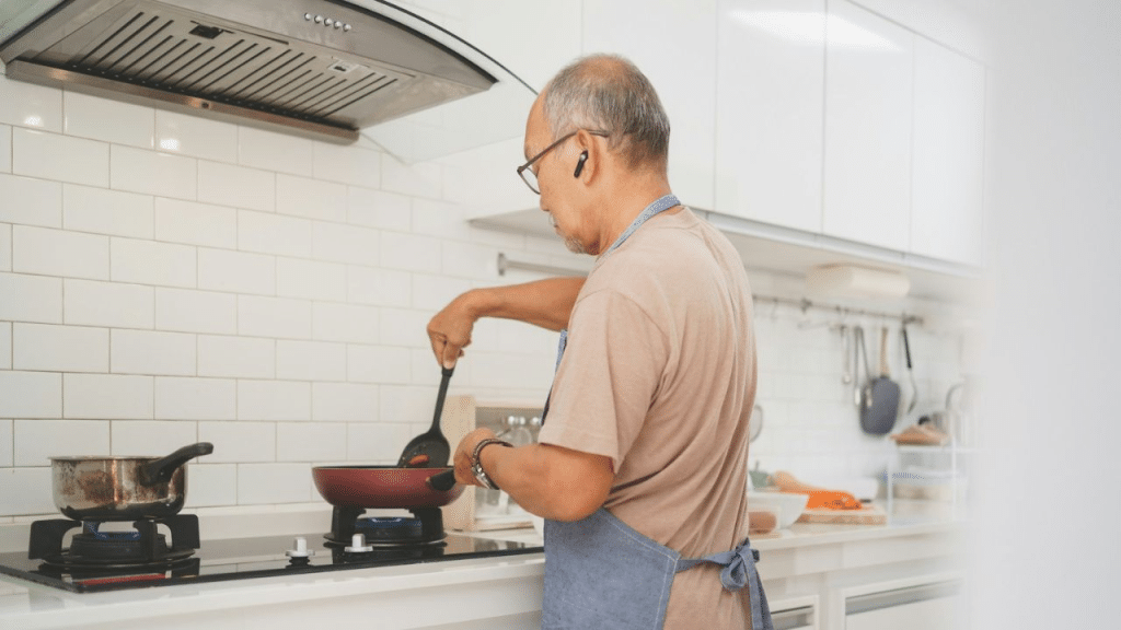 A man with grey hair and glasses is standing in a kitchen and stirring a pan on the stove.