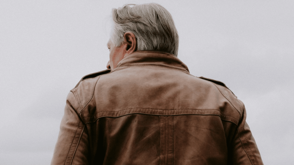 A mature man in a leather jacket is seen from behind, looking away from the camera.