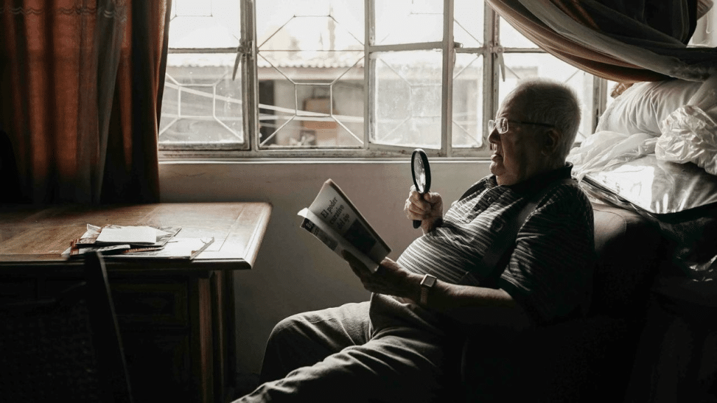 A thoughtful, older man with glasses is seated in a dark room and reading a book with a magnifying glass.