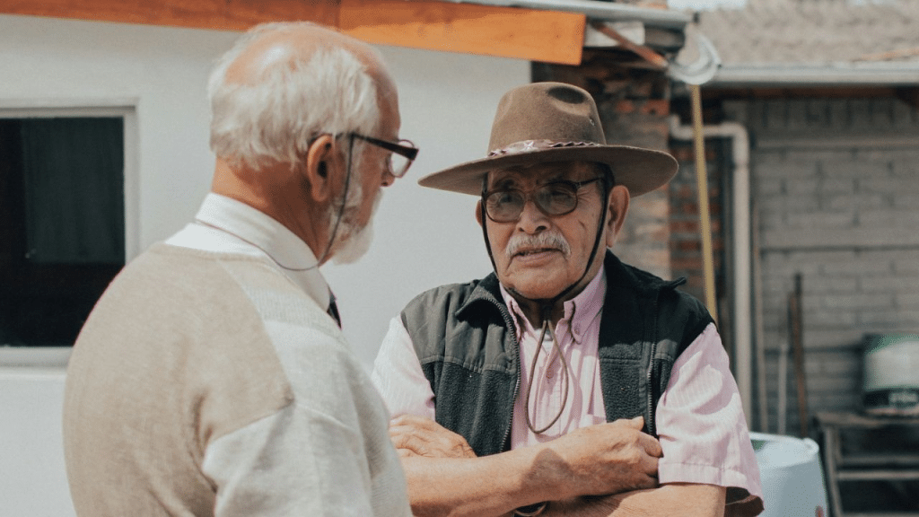 Two senior men, one in a hat, talk outdoors in a sunny setting.