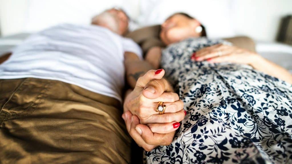 A close-up of an elderly couple lying in bed and holding hands.