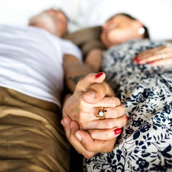A close-up of an elderly couple lying in bed and holding hands.