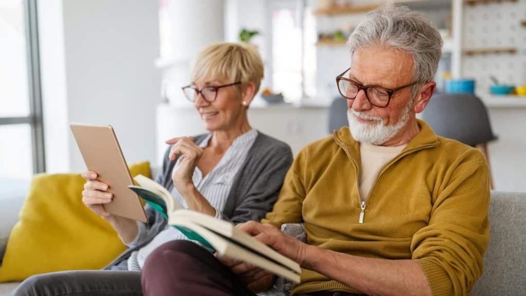 An elderly couple relaxing at home while reading a book and using a tablet.
