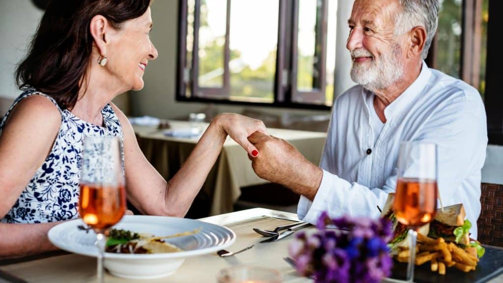 An elderly couple smiling and holding hands while dining at a restaurant.