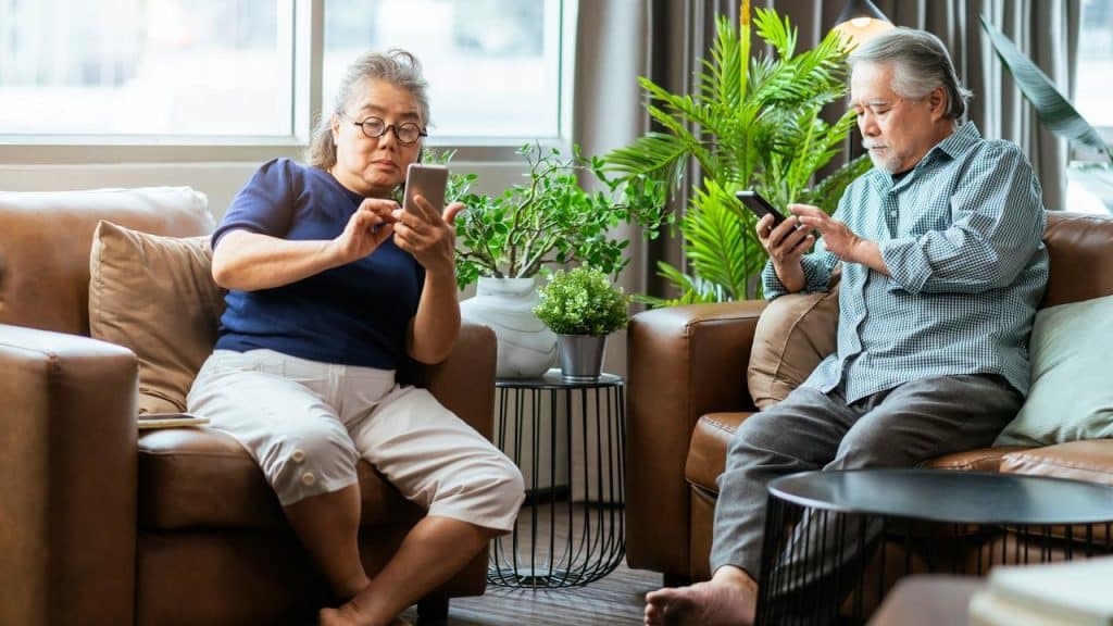 An elderly couple sitting on sofas and using their smartphones.