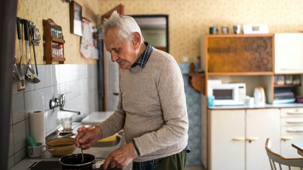 An elderly man with white hair, wearing a sweater, cooking in a kitchen.