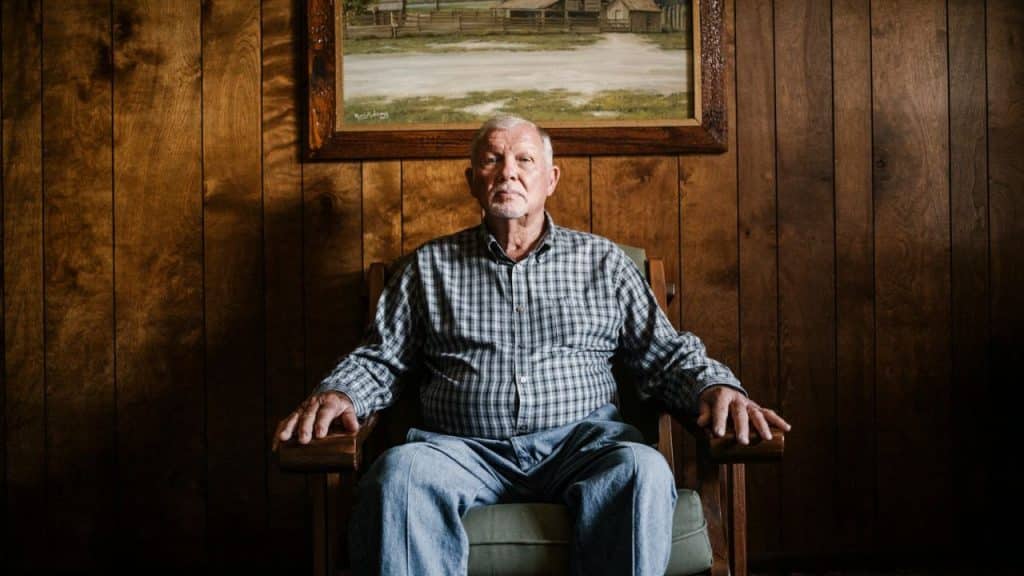 A solemn, older man with a mustache sitting in a wooden chair, facing forward.