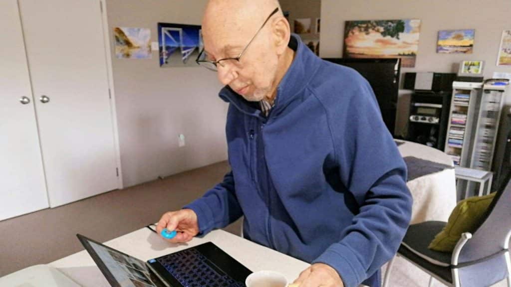 An elderly, bald man with glasses, wearing a blue jacket, uses a laptop.