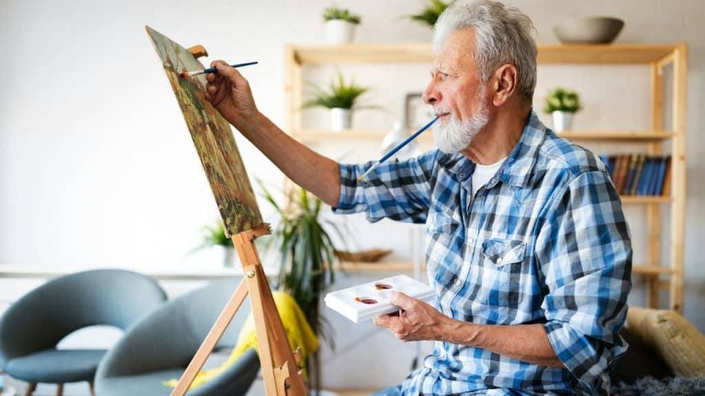 A smiling, older man with a white beard is painting on a canvas.