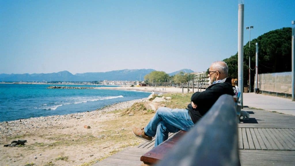 An older man sits on a bench, looking at the ocean and mountains.