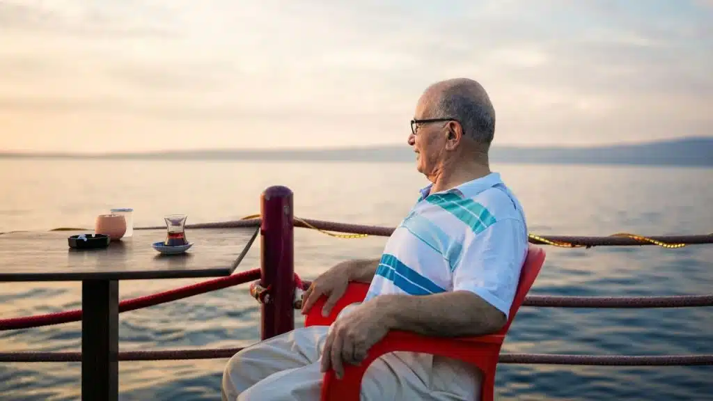 An older man with glasses sits in a red chair overlooking a body of water.