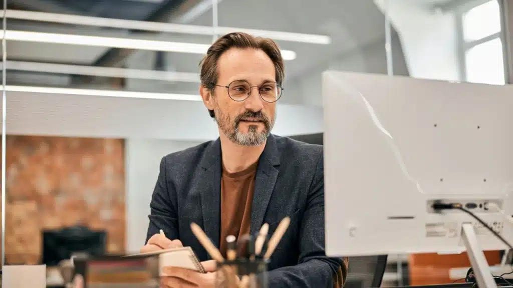 A bearded man in glasses holds a notepad and looks at a computer monitor.