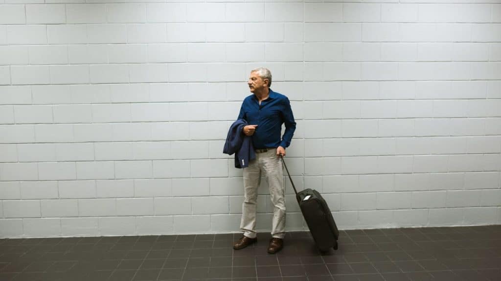 A mature man stands with a suitcase and a jacket in front of a white brick wall.