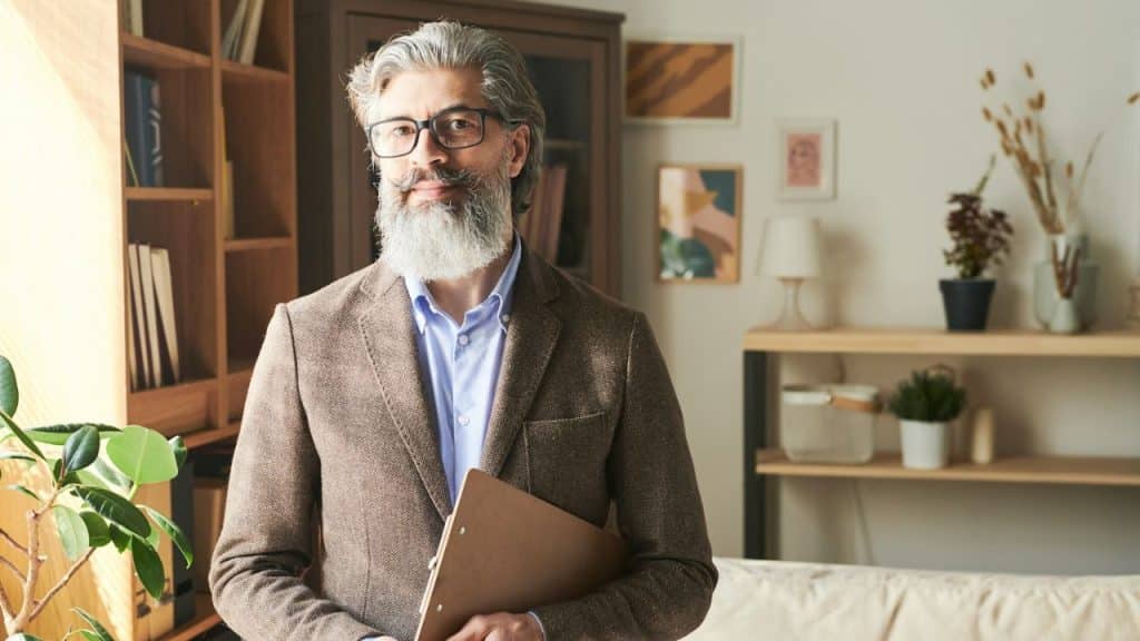 A bearded man in a blazer and glasses holding a clipboard, standing in a room.
