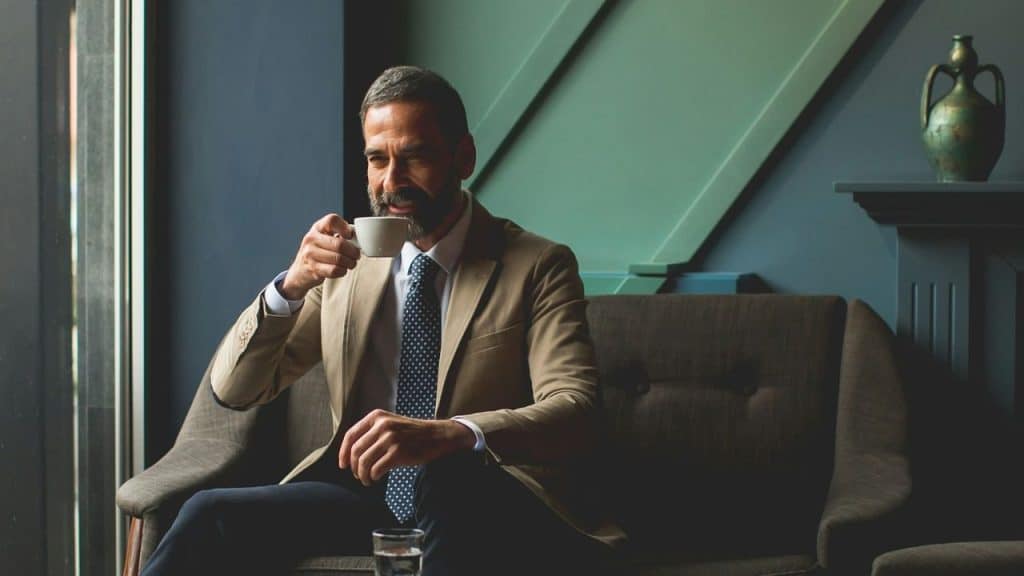A handsome, middle-aged man in a suit sips from a coffee cup while seated.