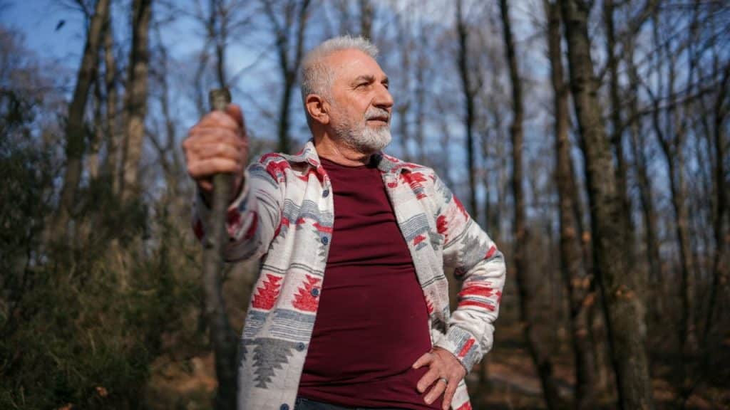 A gray-bearded man in a patterned jacket holds a stick while standing in a forest.