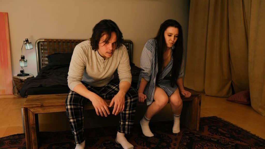 A sad man and woman sit next to each other on a bench in front of a bed.