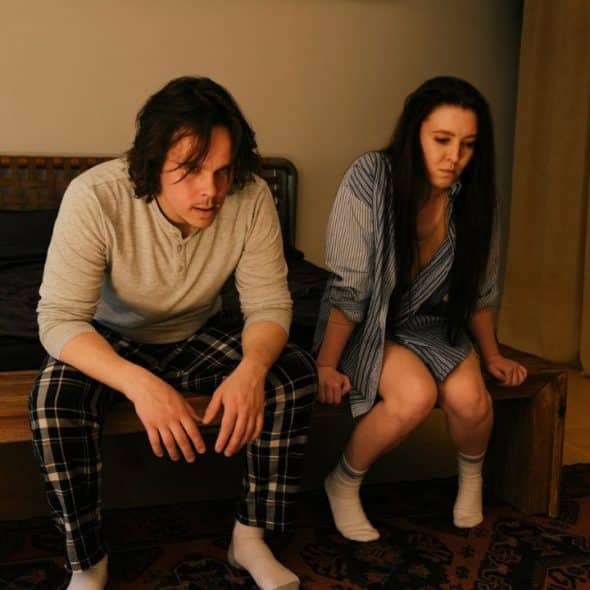 A sad man and woman sit next to each other on a bench in front of a bed.