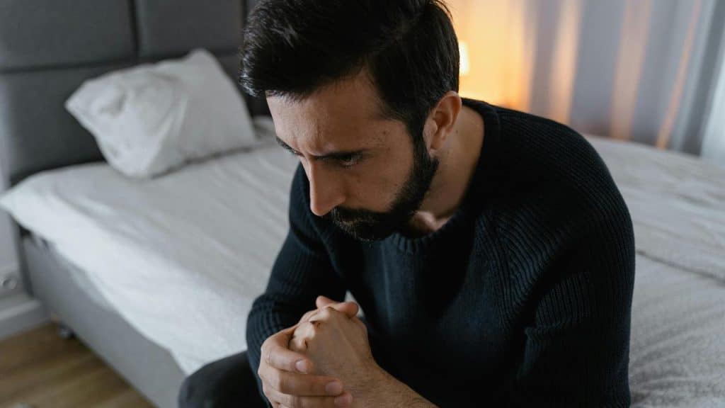 A man sitting on a bed looking pensive with his hands clasped together.