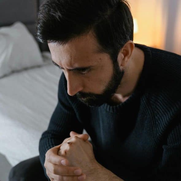 A man sitting on a bed looking pensive with his hands clasped together.