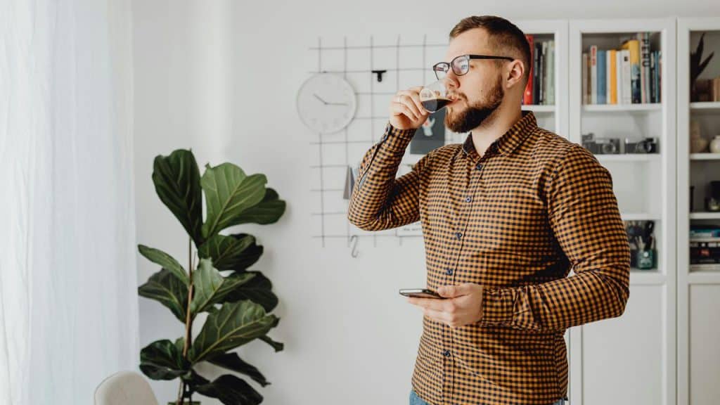 A man standing indoors drinking from a glass while holding a phone.