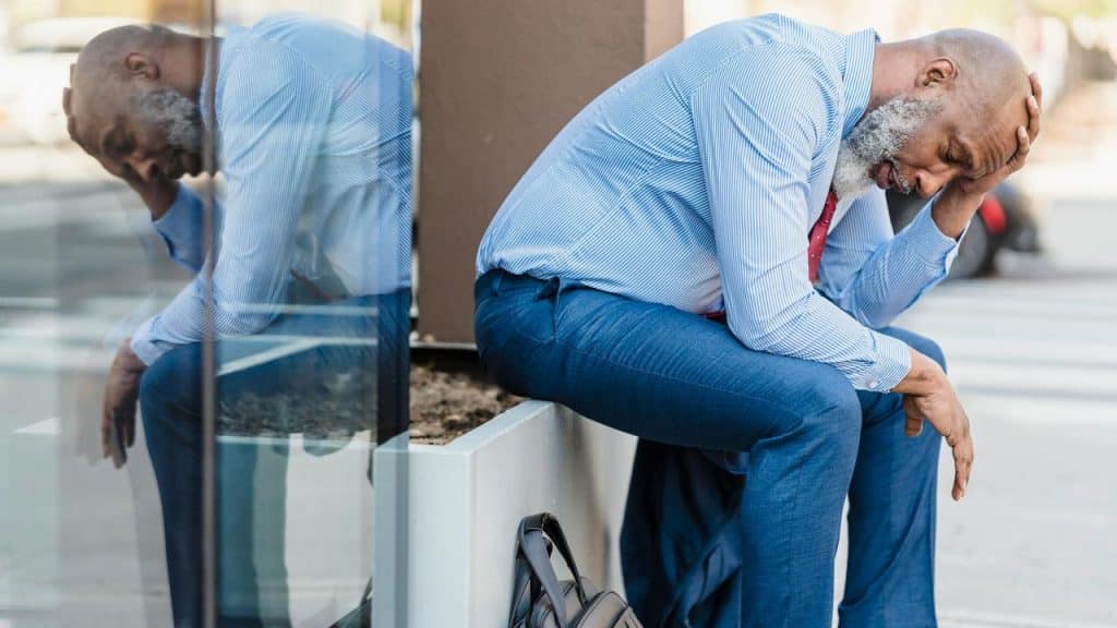 An older man in business attire sitting outside with his head in his hand.