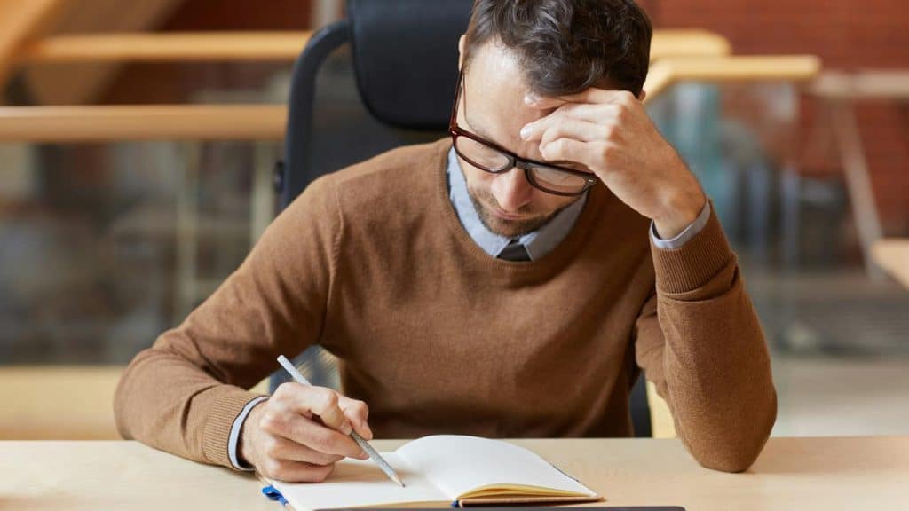A man wearing glasses writing in a notebook while sitting at a desk.