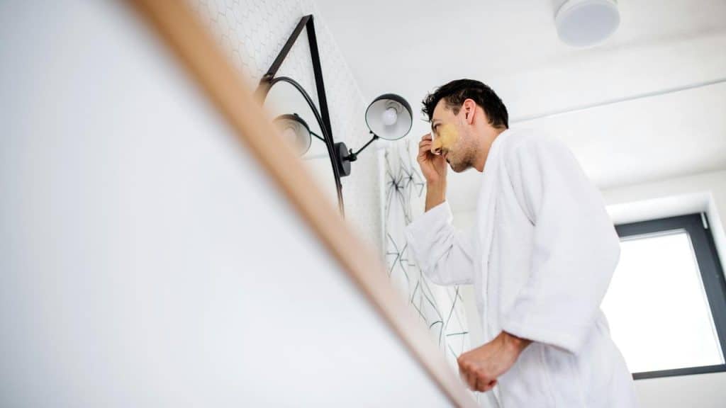 A man in a bathrobe applying a face mask while looking in the mirror.