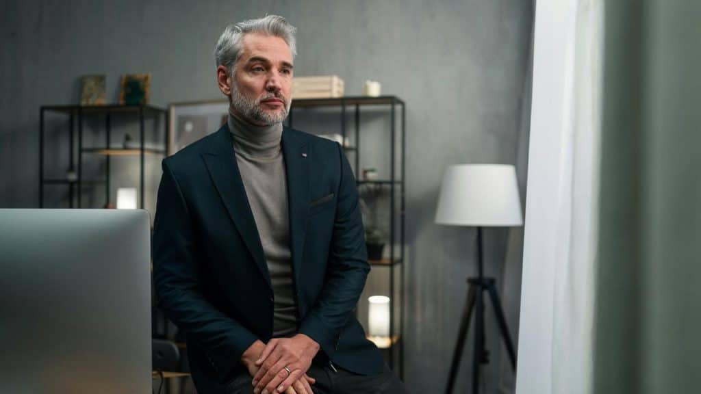 A man with gray hair and a beard wearing a blazer, sitting and looking out the window.