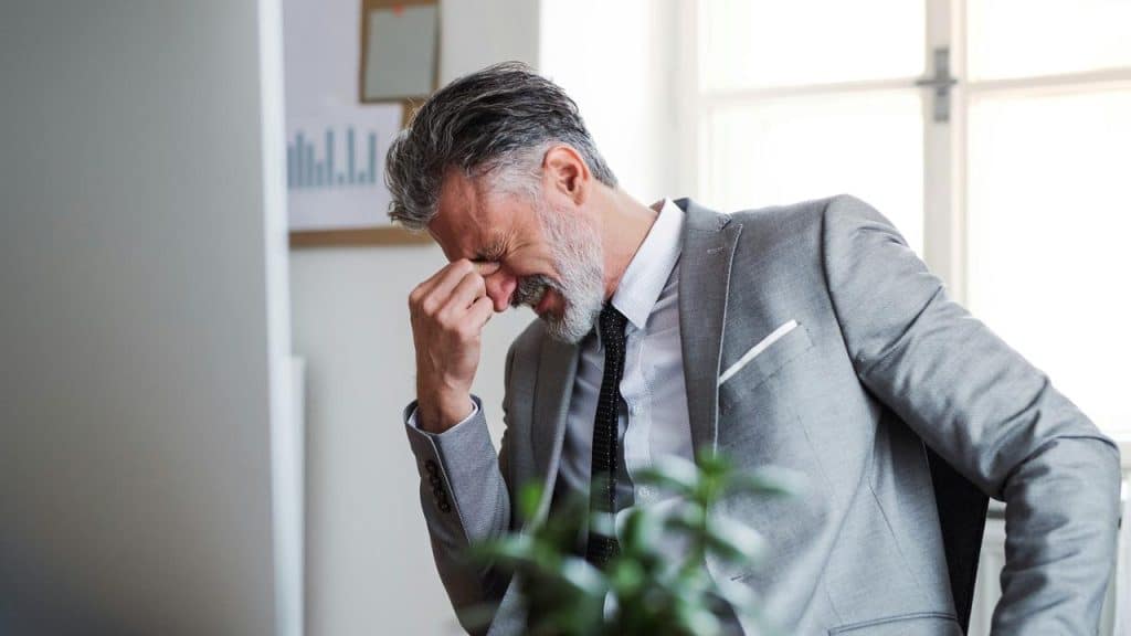A man in a suit sitting at a desk with his hand on his face, looking stressed.