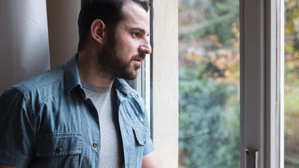 A man standing by a window, looking outside pensively.