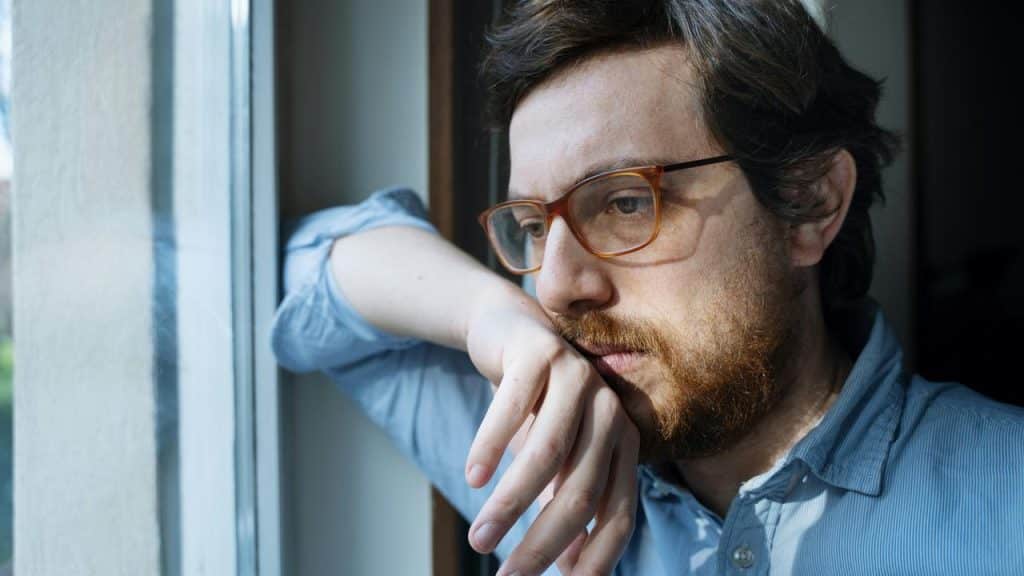 A man with glasses looking out a window thoughtfully with his hand resting on his chin.