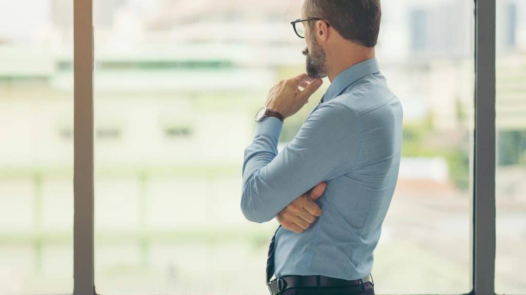 A man in a dress shirt and glasses standing by a window, looking outside thoughtfully.