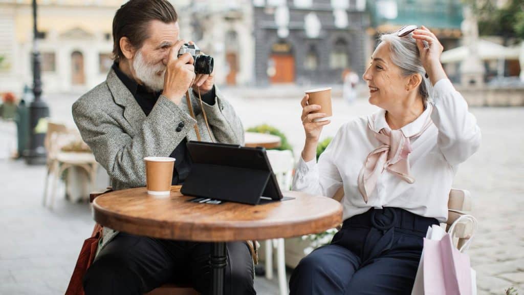 An older couple at a café, with the man taking a photo of the woman.