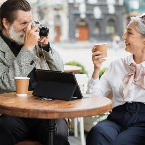 An older couple at a café, with the man taking a photo of the woman.