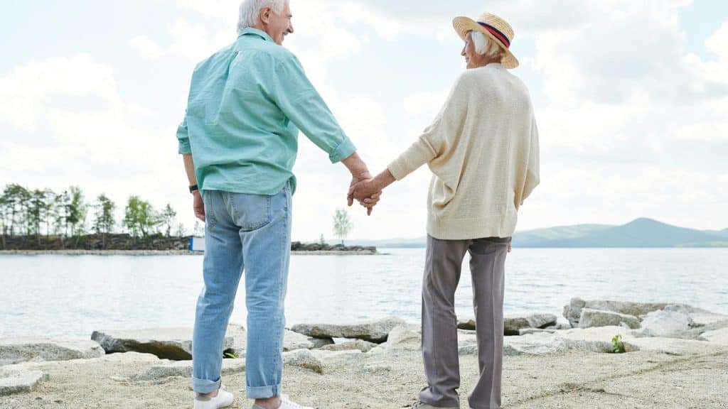 An older couple holding hands while standing by a lake.