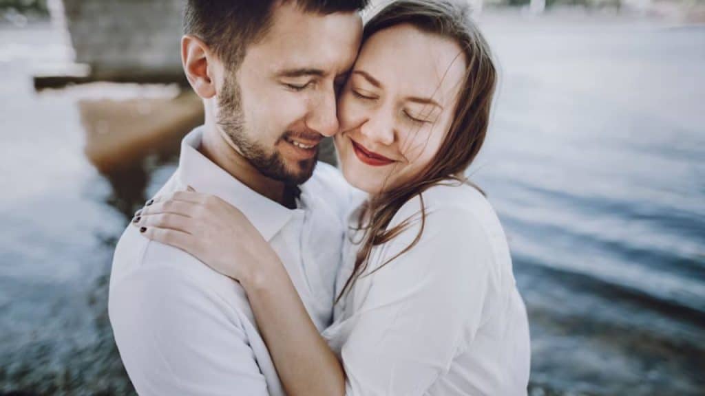 A woman with streaks of gray hair smiling confidently, husband hugging her with admiration