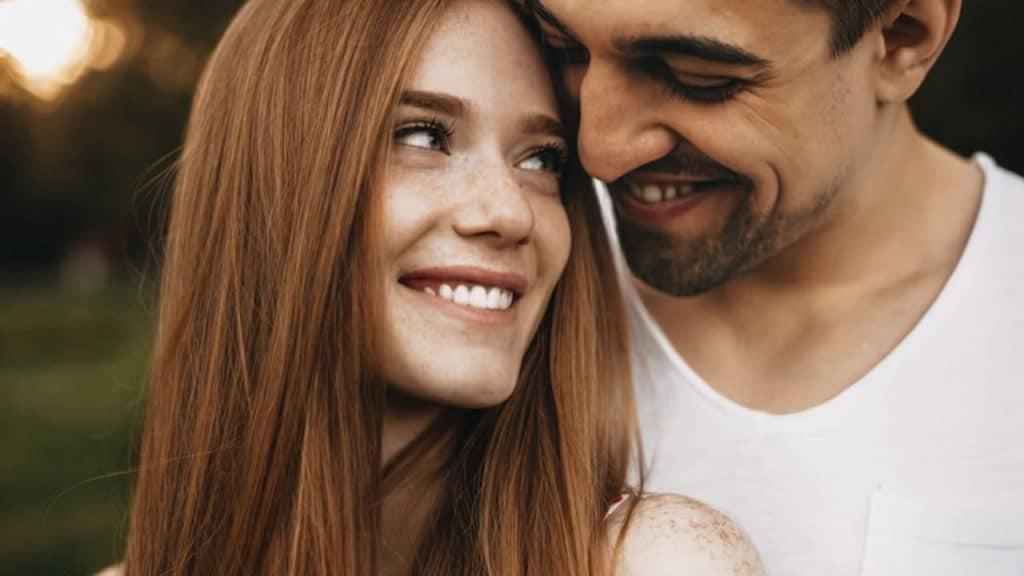 A woman with freckles smiling, her husband hugging her from behind