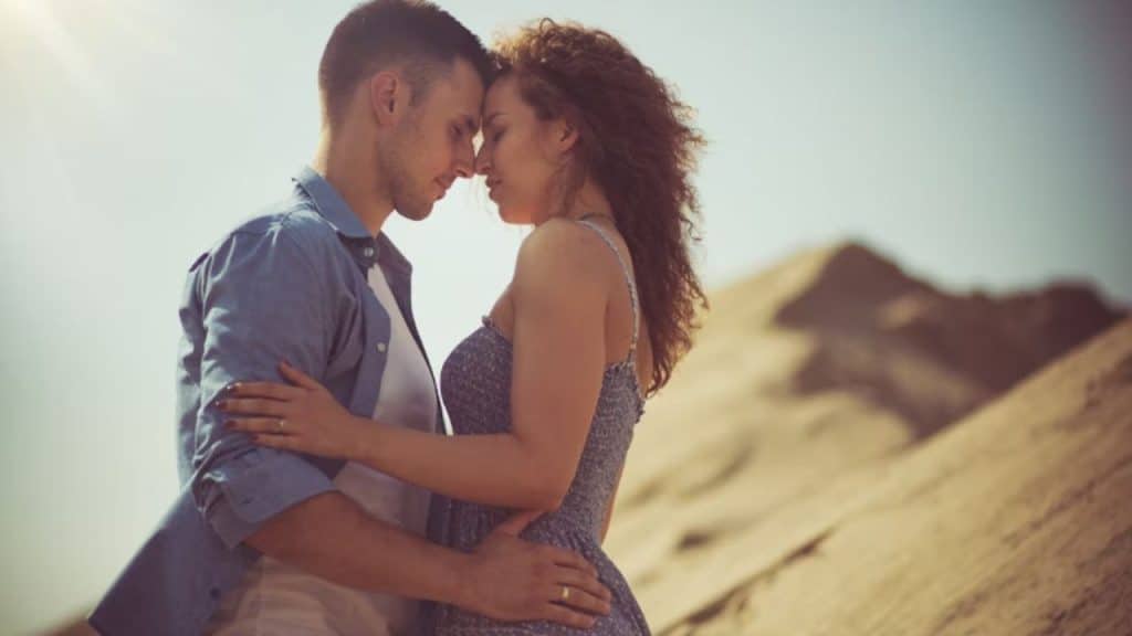 A woman with natural skin texture smiling in sunlight, husband admiring her.