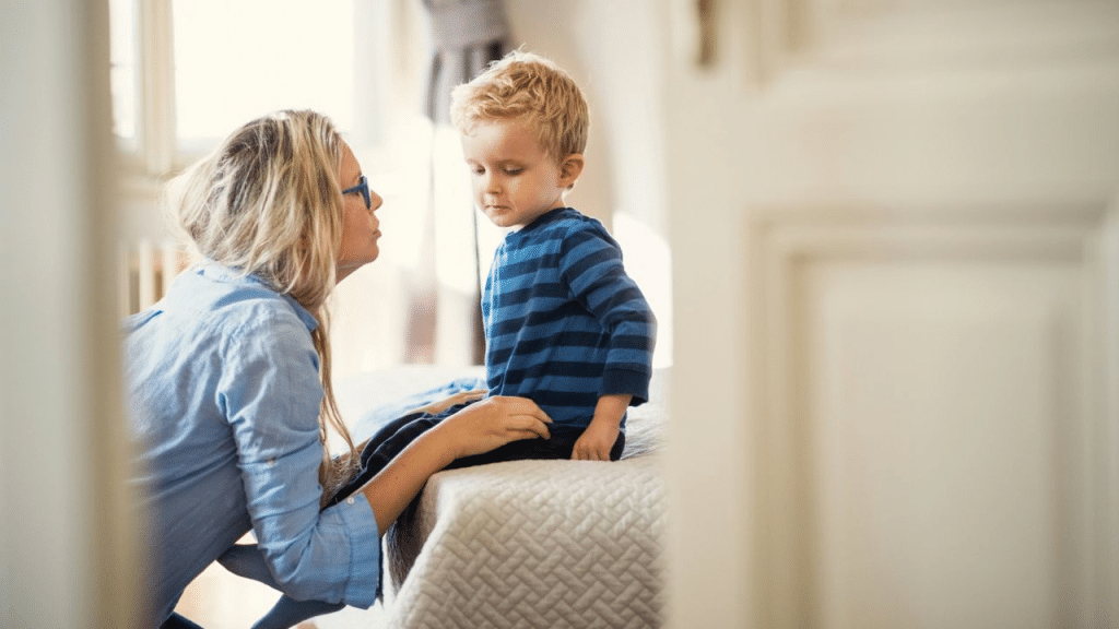 A blonde woman kneels, speaking to a young blonde boy sitting on a bed.
