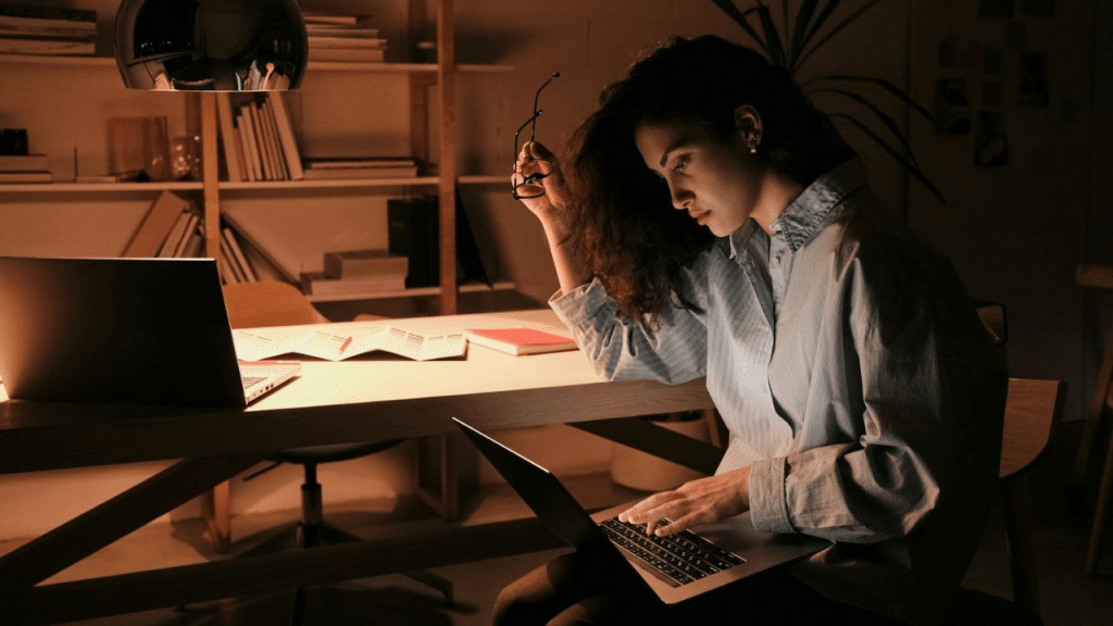 A woman works on her laptop at a desk in a dimly lit room.