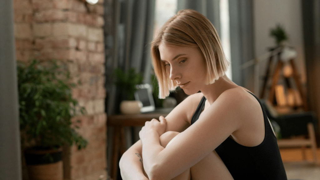 A blonde woman sits with her knees pulled up, looking down with a sad expression.